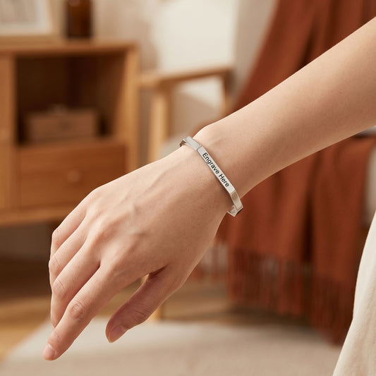 Model shows off small engraved hexagon karha bracelet on wrist in cozy home setting with warm ambient lighting.